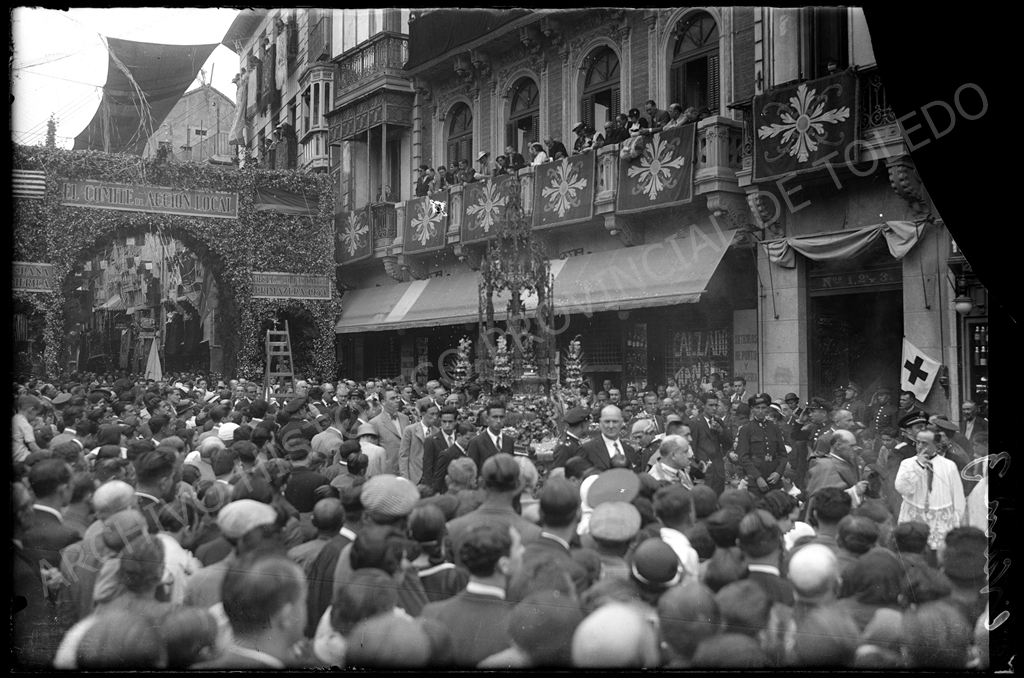 Fotografía de la confluencia de la plaza de Zocodover y la calle Comercio de Toledo. Al principio de dicha calle, un arco cubierto de plantas y con carteles. Una gran multitud y, en el centro de la imagen, la custodia, rodeada de varios hombres, tanto civiles como militares.
